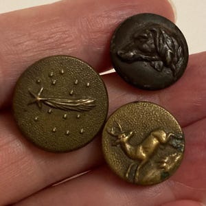 May include: Three antique brass buttons with detailed designs. One button features a shooting star, another a dog's head, and the third a leaping deer. The buttons have a vintage, worn appearance, suggesting they are old or have been well-used.