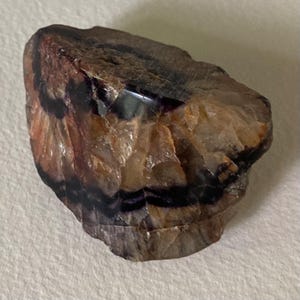 Può includere: Una gemma grezza e non tagliata con un mix di colori. La pietra presenta bande di viola scuro e nero su uno sfondo di tonalità beige, marrone e arancione. La superficie ha una consistenza ruvida e naturale.