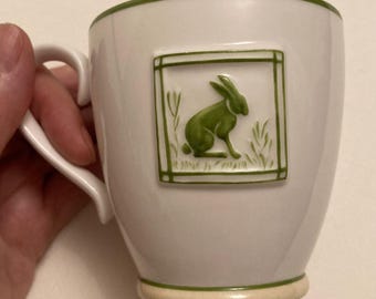 Może przedstawiać: Biały ceramiczny kubek z zieloną obwódką i uchwytem. Zielony wzór królika znajduje się w prostokątnej ramce z przodu. Kubek ma beżową podstawę i jest trzymany przez dłoń.