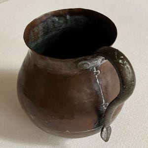 May include: A copper-colored, round-bodied pot with a dark interior and a curved handle. The pot has a wide opening and a riveted handle. The metal shows signs of age and wear, with some white discoloration.
