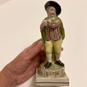 Puede incluir: Una figura de porcelana vintage de un hombre con atuendo del siglo XVIII. La figura lleva un sombrero negro, una capa verde y un chaleco estampado con calzones amarillos. La figura está montada sobre una base rectangular con un diseño pintado.