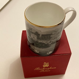 May include: White ceramic mug with a gold rim, featuring a black and white image of a building and swans. The mug sits on a red box with the words "Buckingham Palace" in gold lettering.