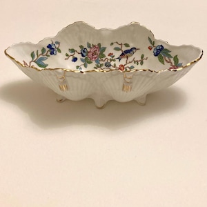 May include: A white porcelain serving dish with a scalloped edge and three small feet. The dish is decorated with a floral and bird design in blue, pink, and green, with gold trim. A decorative piece for serving.