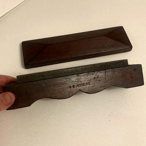 Könnte beinhalten: Ein dunkelbraunes Holzwerkzeug mit rechteckiger Form und Metallklinge. Das Werkzeug hat eine gebogene Kante und der Text "G.E. ROGERS" ist sichtbar. Das Oberteil ist ein dunkelbrauner Holzdeckel.