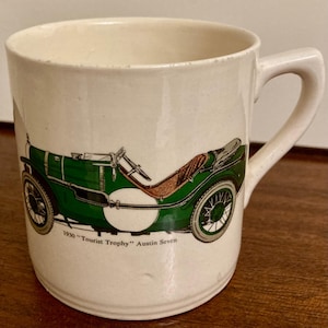 Puede incluir: Taza de cerámica color crema con una ilustración de un coche antiguo verde. El coche tiene asientos marrones y ruedas negras. El texto en la taza dice "1930 'Tourist Trophy' Austin Seven". La taza tiene un asa curva.