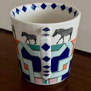 Puede incluir: Taza de cer&aacute;mica blanca con un patr&oacute;n geom&eacute;trico en azul, naranja y verde azulado. La taza presenta ilustraciones de cebras en blanco y negro y un asa con detalles de diamantes azules. El borde tiene un borde de diamantes azules.