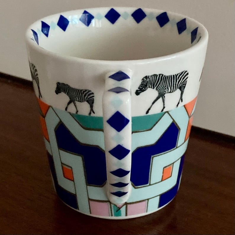 Puede incluir: Taza de cer&aacute;mica blanca con un patr&oacute;n geom&eacute;trico en azul, naranja y verde azulado. La taza presenta ilustraciones de cebras en blanco y negro y un asa con detalles de diamantes azules. El borde tiene un borde de diamantes azules.