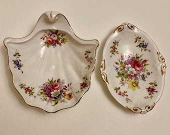 May include: Two white porcelain dishes with floral designs. One is shell-shaped with a gold trim, and the other is oval with a scalloped gold edge. Both feature bouquets of pink, red, and yellow flowers with green leaves.