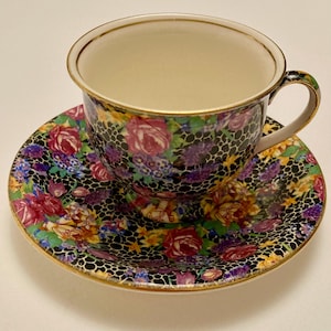 Puede incluir: Un juego de taza y platillo de té vintage. La taza y el platillo presentan un fondo negro con un estampado floral en tonos rosas, morados, amarillos y verdes. Los bordes están adornados con dorado.