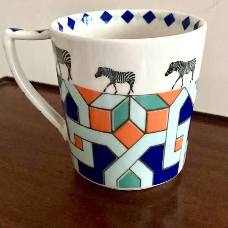 Puede incluir: Taza de cer&aacute;mica blanca con un patr&oacute;n geom&eacute;trico en azul, naranja y verde azulado. La taza presenta tres ilustraciones de cebras y un asa. El borde est&aacute; decorado con formas de diamante azules.