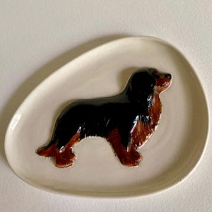 May include: A ceramic decorative plate with a sculpted Cavalier King Charles Spaniel dog. The dog is black and brown, and the plate is off-white. The plate has a smooth, glossy finish and is oval-shaped.