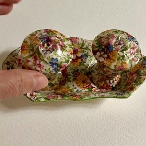 Pode incluir: Saleiro e pimenteiro florais com uma bandeja a condizer. O conjunto apresenta um padrão floral colorido com flores rosa, amarelas, azuis e verdes. A bandeja tem uma borda ondulada.