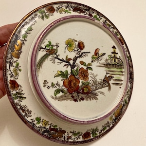 May include: A decorative ceramic plate with an Asian-inspired design. The plate features a central scene with birds, flowers, and a pagoda, framed by a border of floral and gold accents. The plate has a diameter of approximately 20 cm.