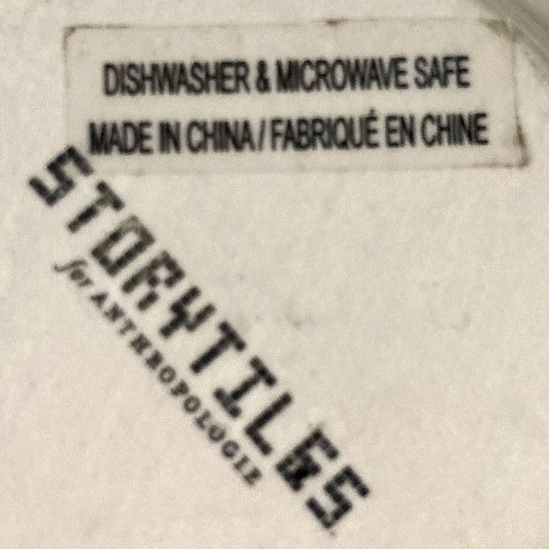 Puede incluir: Primer plano de una etiqueta blanca con texto negro. La etiqueta dice "Dishwasher & Microwave Safe" y "Made in China / Fabriqu&eacute; en Chine". Debajo, la palabra "Storytiles" est&aacute; impresa diagonalmente en negro, con "for Anthropologie" debajo.