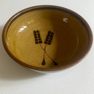 Puede incluir: Un cuenco redondo de cerámica con un diseño de espigas de trigo. El cuenco tiene un interior marrón claro y un borde marrón más oscuro. Dos espigas de trigo negras están pintadas en el centro del cuenco.