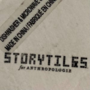 Puede incluir: Primer plano de una superficie blanca con el texto "STORYTILES for ANTHROPOLOGIE" en negro. Es visible una etiqueta con el texto "DISHWASHER & MICROWAVE SAFE MADE IN CHINA / FABRIQU&Eacute; EN CHINE".