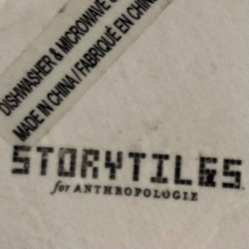 Puede incluir: Primer plano de una superficie blanca con el texto "STORYTILES for ANTHROPOLOGIE" en negro. Es visible una etiqueta con el texto "DISHWASHER & MICROWAVE SAFE MADE IN CHINA / FABRIQU&Eacute; EN CHINE".