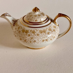 Op de afbeelding: Een kleine, witte porseleinen theepot met gouden details. De theepot heeft een gouden handvat, tuit en dekselknop. De body is versierd met een gouden bloemmotief. Gouden banden sieren het midden en de basis.
