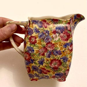 Puede incluir: Una jarra de crema floral vintage con un fondo amarillo y un patrón de flores rojas, azules y moradas. La jarra tiene un asa y un pico, y el borde está adornado con oro.