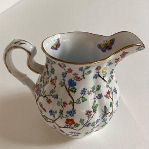 May include: White ceramic jug with a gold rim and handle. The jug is decorated with colourful butterflies and floral branches in red, blue, green, and yellow. The handle has a bamboo-like design.