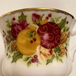 以下が含まれることがあります： 金色の縁取りが付いた白いティーカップ。クリスマスのオーナメントのデザインが施されています。デザインには、金のオーナメント、赤いオーナメント、赤いベリー、緑の葉、黄色と赤のバラが含まれています。