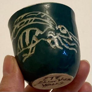 Puede incluir: Una pequeña taza de cerámica color teal hecha a mano con un diseño abstracto blanco. La taza tiene forma redondeada y un acabado brillante. La base de la taza tiene el texto "ETE Vale Ebbw Wales".