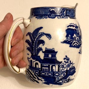 May include: White ceramic pitcher with a blue Willow pattern design. The pitcher has a curved handle with gold trim and a spout. The blue design features pagodas, trees, and a boat scene. The rim of the pitcher has a blue patterned border.