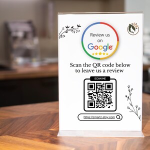 Peut inclure: Panneau en acrylique blanc avec le logo Google et le texte « Évaluez-nous sur Google ». Il comprend également un code QR et le texte « Scannez le code QR ci-dessous pour nous laisser un avis ». Le panneau est sur une surface en bois.