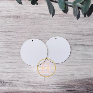 May include: Two white, round wooden ornaments with a black edge and a small hole at the top for hanging. The ornaments are blank, ready for customization. The background is a light wood grain.