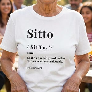 May include: A white t-shirt with the word "Sitto" in large black letters. Below the word is the definition of "Sitto", which is "Like a normal grandmother but so much cooler and Arabic."