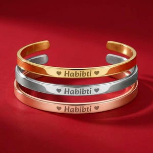May include: Three open cuff bracelets in gold, silver, and rose gold. Each bracelet is engraved with the word "Habibti" between two heart symbols. The bracelets are displayed on a red surface.