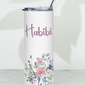 May include: White tumbler with a floral design and the word "Habibi" in purple script. The floral arrangement includes pink, blue, and white flowers with green leaves. A silver straw is inserted into the lid.
