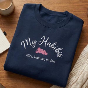 May include: Navy blue sweatshirt with the text "My Habibis" in white script, along with three pink hearts. Below the hearts are the names "Alice, Thomas, Jordan" in a smaller font.