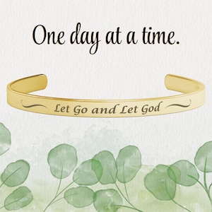 May include: Gold-toned bangle bracelet with the inscription "Let Go and Let God" on a white background with green foliage.
