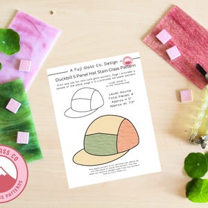 May include: A flat lay featuring a stained glass pattern design for a duckbill hat. The design is printed on a white sheet of paper, with the Fuji Glass Co. logo. The image includes pink and green glass pieces, and a glass cutter.
