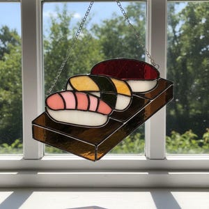 May include: Stained glass sushi art piece featuring three sushi pieces on a brown rectangular base. The sushi includes a piece with pink and white elements, a yellow and green piece, and a red and white piece. A silver chain is attached for hanging.