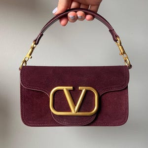 May include: A deep burgundy suede handbag featuring a rectangular silhouette with a flap closure. The front is adorned with a large, matte gold-toned metal V logo emblem. It includes a matching burgundy suede top handle attached with gold-toned hardware.