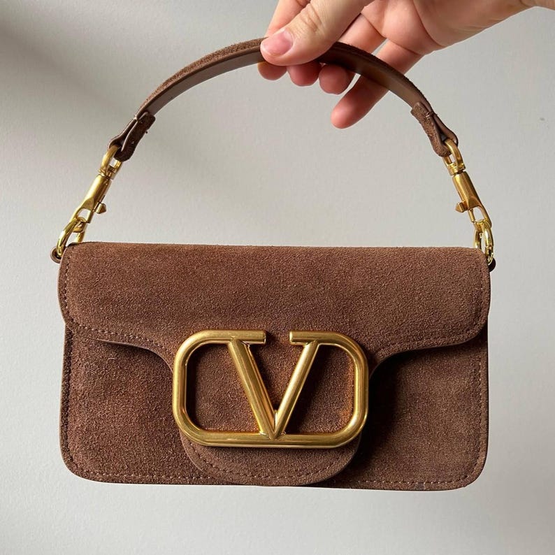 以下が含まれることがあります： 前面に金色の金属製「V」ロゴが付いた茶色のスエードハンドバッグ。バッグにはお揃いの茶色のハンドルと金色の金具が付いています。長方形のバッグにはフラップクロージャーが付いています。