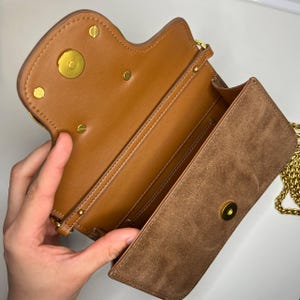 以下が含まれることがあります： 茶色の革製クロスボディバッグのオープンビュー。スエードのフラップが付いています。ゴールドチェーンのストラップとゴールドの金具が特徴です。内側は茶色の革で裏打ちされ、複数のコンパートメントがあります。サドルバッグの形にデザインされています。