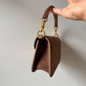 以下が含まれることがあります： 長方形の形とフラップクロージャーを備えた茶色のスエードハンドバッグ。バッグは短いハンドルと金色のハードウェアを備えています。ハンドルは金色の留め金でバッグに取り付けられています。バッグは手で持たれています。