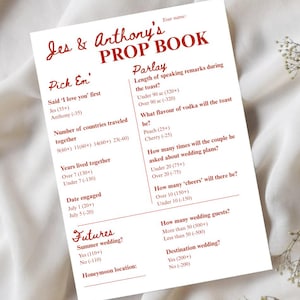 May include: A white paper titled "Jes & Anthony's PROP BOOK" with red text. The book contains wedding game questions like "How many wedding guests?" and "Summer wedding?" with corresponding point values. The paper is on a white fabric.