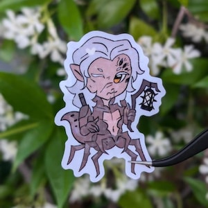 May include: A sticker depicting a fantasy character with white hair, pointed ears, and multiple eyes. The character has a spider-like body and holds a lantern. The sticker is in a cartoon style with brown and tan colours.
