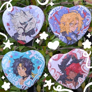 May include: Four heart-shaped badges featuring anime-style character illustrations. Each badge showcases a different character with distinct hair and clothing, set against colourful backgrounds with decorative elements. The badges are displayed against a backdrop of green foliage.