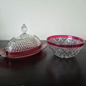 May include: A clear glass butter dish with a diamond-patterned dome lid and a red glass base, accompanied by a matching clear glass bowl featuring a diamond-cut exterior and a vibrant red rim, both displaying a reflective and textured surface.