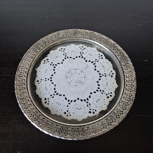 May include: A round glass plate featuring a white paper doily centred on its surface. The plate is framed by a wide, metallic silver rim with an intricate, openwork filigree pattern that runs along the entire outer edge of the circular dish.