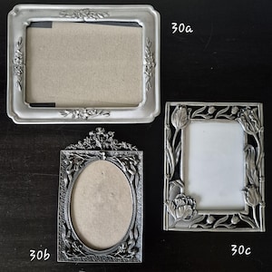 May include: Three decorative metal picture frames. The frames are silver-toned and feature floral and botanical designs. One frame is rectangular with rounded corners, another is oval, and the third is square with tulip motifs.