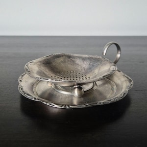 May include: A silver-toned metal tea strainer resting on a matching circular drip tray. The strainer features a shallow bowl with a perforated base, a curved handle, and small spherical feet. The tray has a decorative scalloped edge.