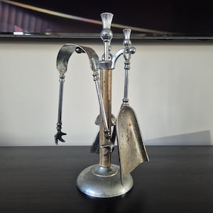 May include: A silver-toned metal fireplace tool set featuring a central stand with a round base, a shovel, and a pair of tongs with claw-like ends. The central column has a light brown wooden accent, and the handles are topped with thistle-shaped finials.
