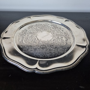 May include: A round silver-toned metal serving tray with a scalloped edge and a wide, smooth rim. The centre of the tray features an intricate, embossed floral and scrollwork pattern that covers the circular surface, creating a textured design.
