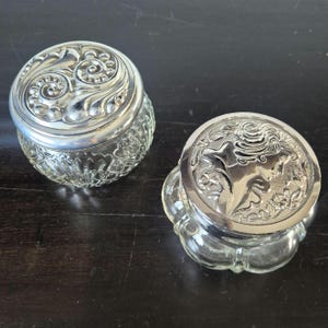 May include: Two small glass jars with silver-toned metal lids. The left jar features a clear glass body with a diamond-cut pattern and a lid with swirling embossed designs. The right jar has a smooth, ribbed glass base and a lid depicting a profile.